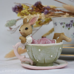 Rabbit in pink and white dress in a green teacup with pale pink saucer. Rabbits arms resting on the cup handle