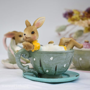 Rabbit in yellow and white dress in a grey green teacup. Rabbit leaning on cup handle