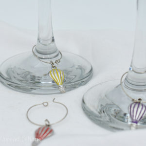 wine glass charms of hot air balloons in red, yellow and purple. the yellow are purple are attached to glasses, the red on on a white tablecloth