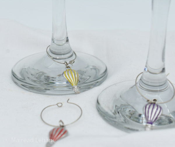 wine glass charms of hot air balloons in red, yellow and purple. the yellow are purple are attached to glasses, the red on on a white tablecloth