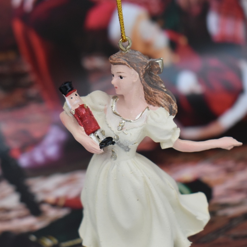 ballerina with nutcraker doll in her arms with out stretched arm , wearing a pale cream dress with gold trim and her long hair tied back in a bow